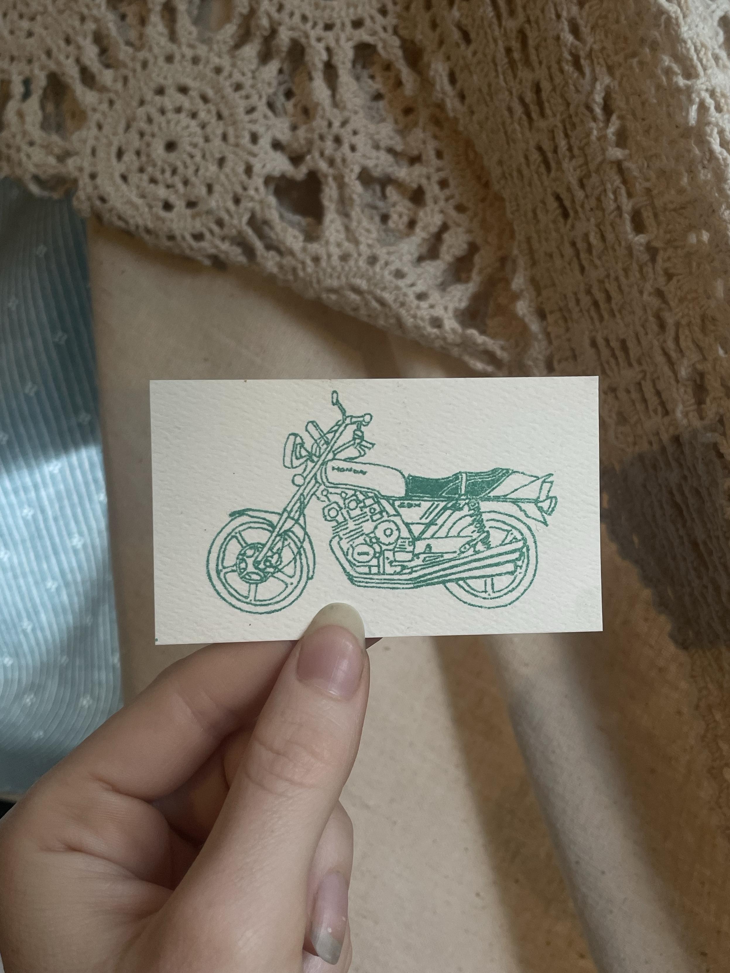 An illustration of a motorcycle is printed on a small, textured card being held by a hand. The design is in seafoam ink, and the background features a crochet fabric and textured surfaces.